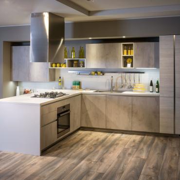 Graue L-Küche in Zementoptik mit sichtbaren Wänden und Regalen, Deckenabzugshaube, Kühlschrank und ausziehbarem Vorratsschrank